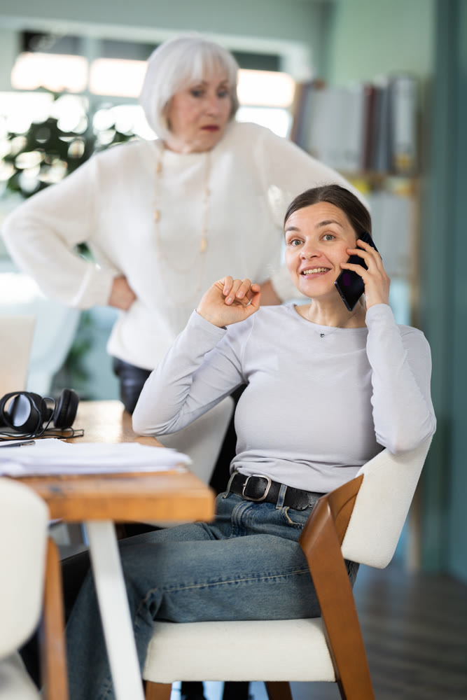 Elderly woman judging female colleague talking on mobile phone
