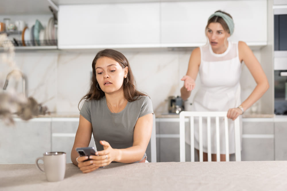 Young girl sitting at the table while woman is quarreling with her standing behind