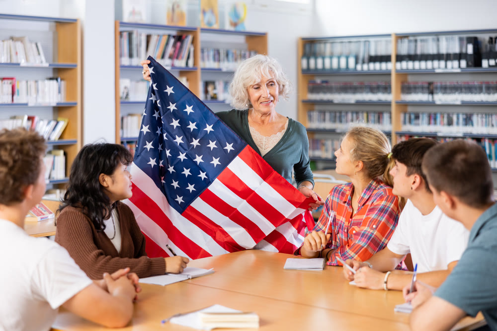 Mature female teacher shows the flag of United States of America to teenagers and talks about this country during lesson at library school