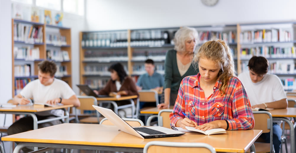 Focused teenager female student sitting at library desk, preparing for exam