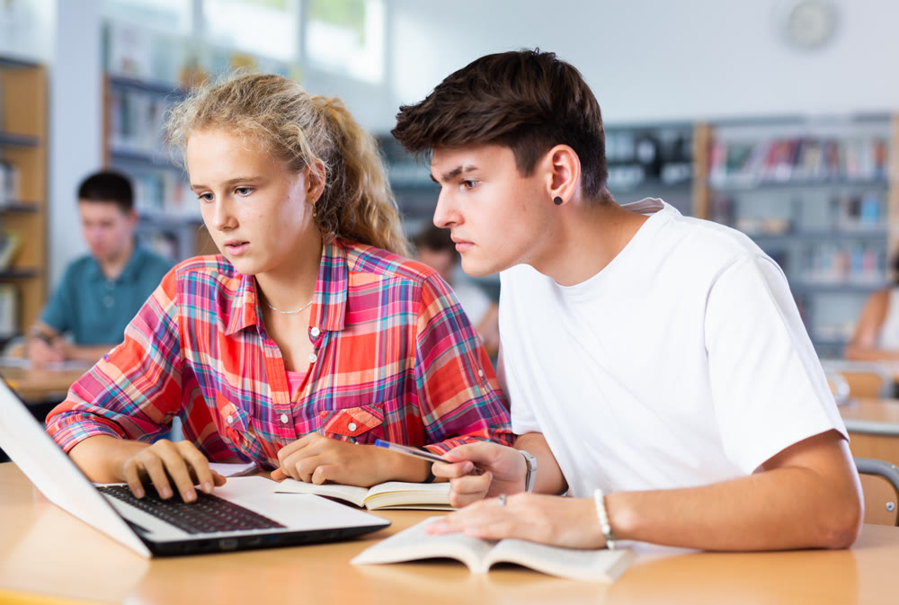 Positive female and male students teenagers study together at laptop while sitting in school library