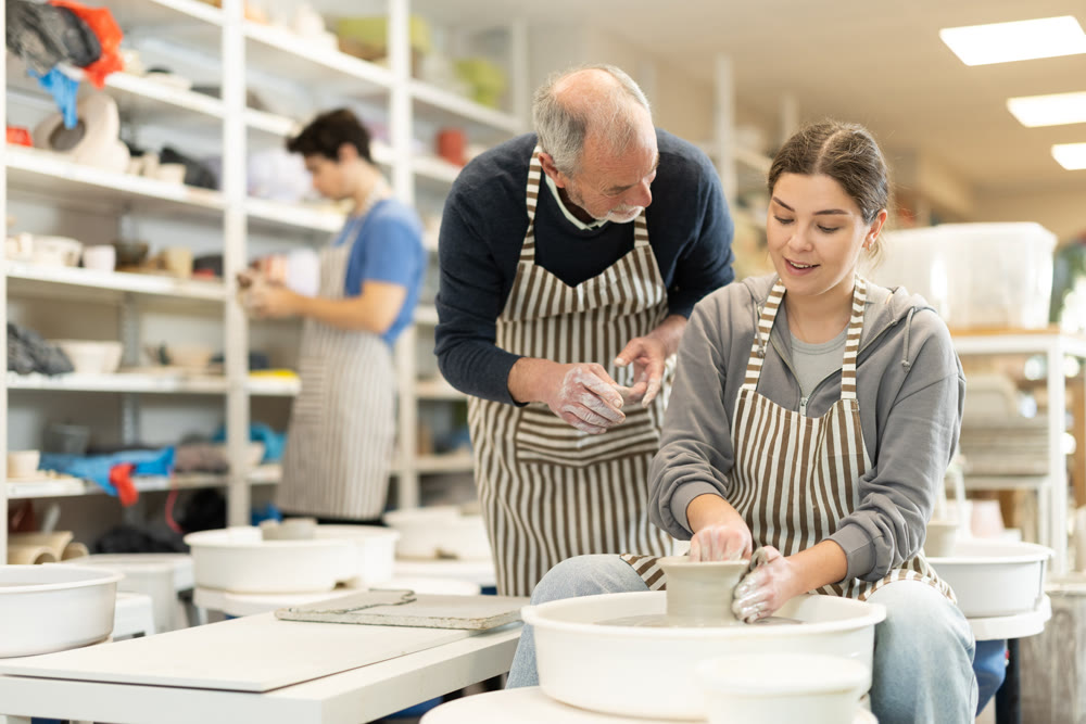 Mature man help to girl working on potters wheel in pottery workshop