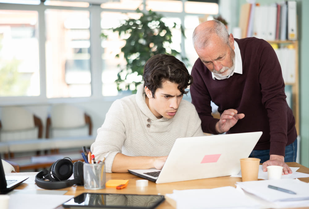 Elderly man helping young male colleague work on laptop
