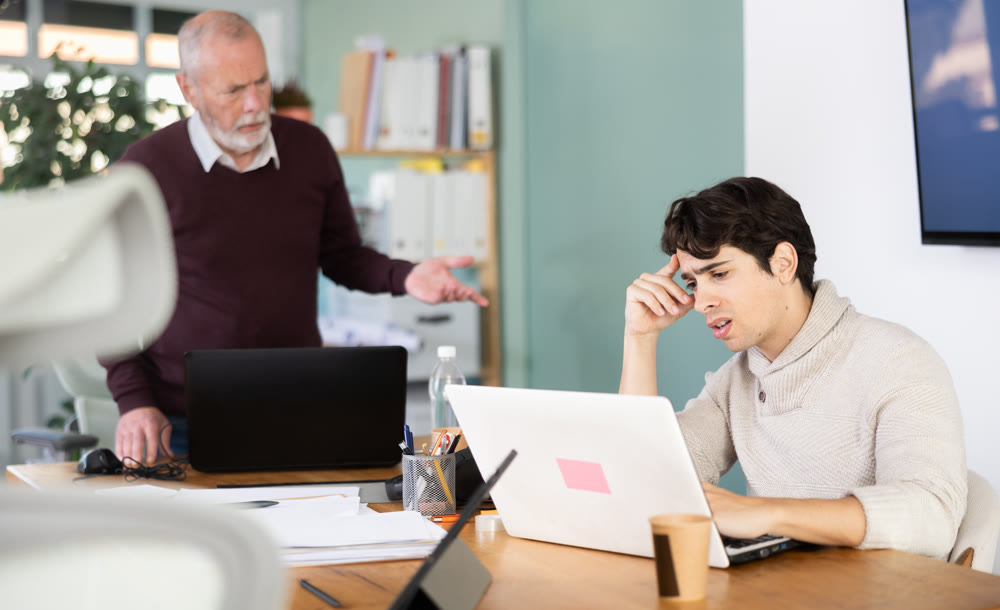 Senior man head of department shouts at guy subordinate who is working at laptop