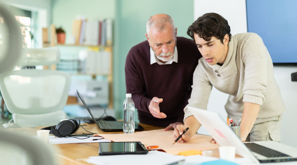 Two men are bent over drawing, reviewing and discussing development of construction.