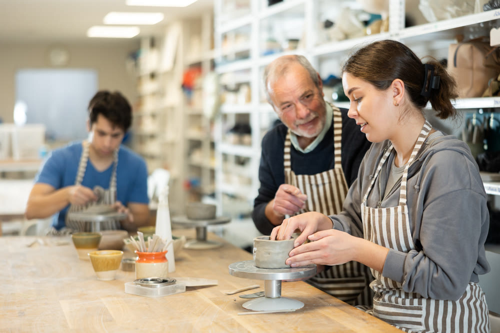 Elderly man teacher shows girl how to sculpt ceramic product from raw clay in workshop