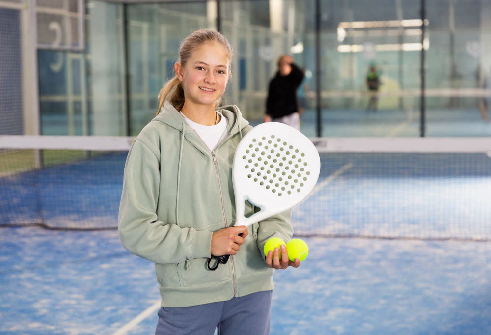 Positive teenage girl looking at camera while playing padel at court