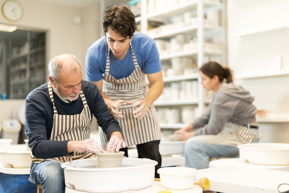 Young guy helping elderly man shape pottery on wheel