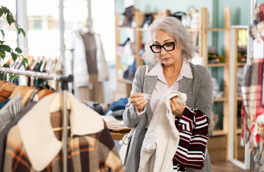 Mature female buyer in glasses chooses fashionable sweater in clothing store