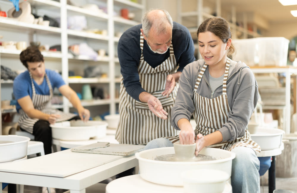 Eldery man helping young woman shape pottery on wheel