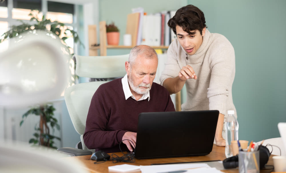 Young man helping elderly male colleague work on laptop