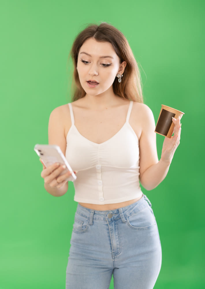 Woman standing on green background with smartphone and coffee in hands