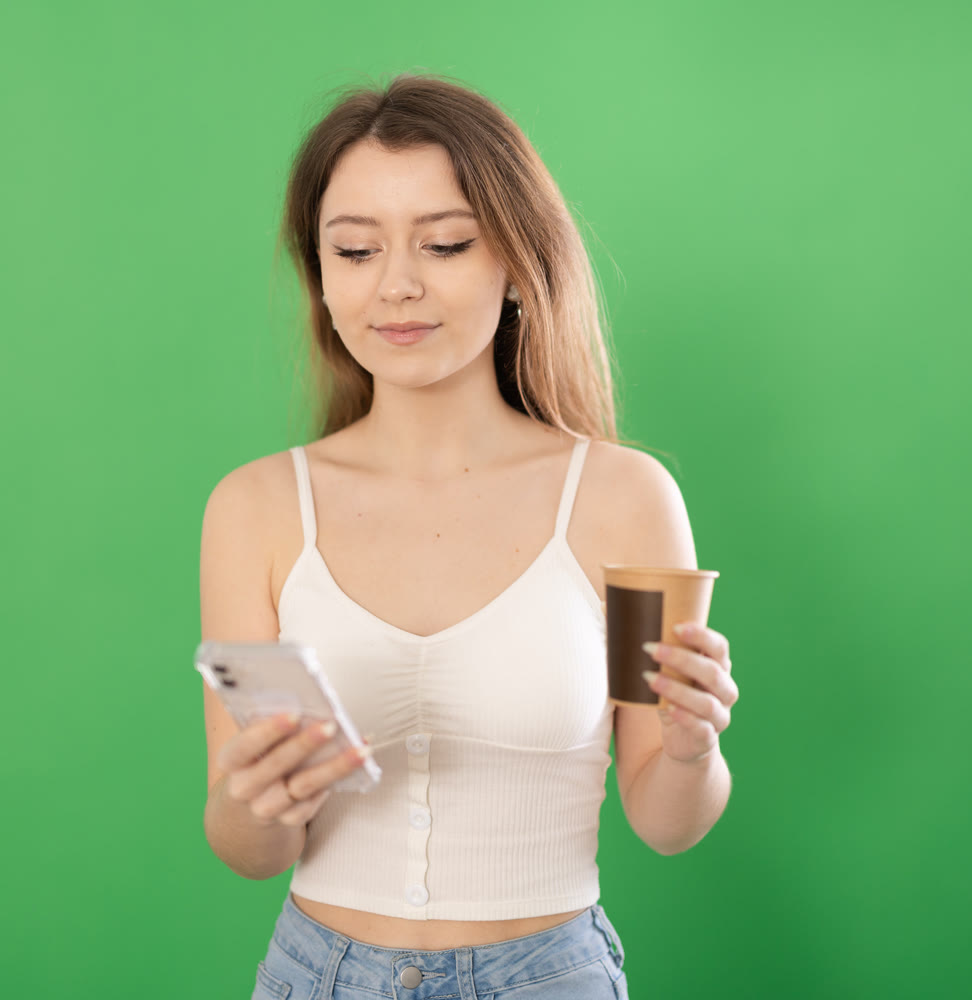 Woman standing on green background with smartphone and coffee in hands
