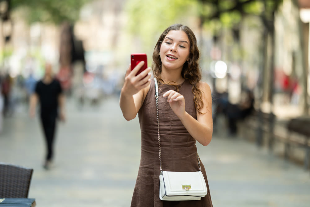 Beautiful stylish young girl in dress with white handbag walking and posing on city street on summer day and taking selfies with mobile phone
