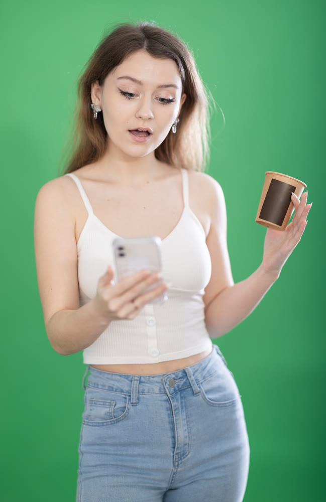Woman standing on green background with smartphone and coffee in hands