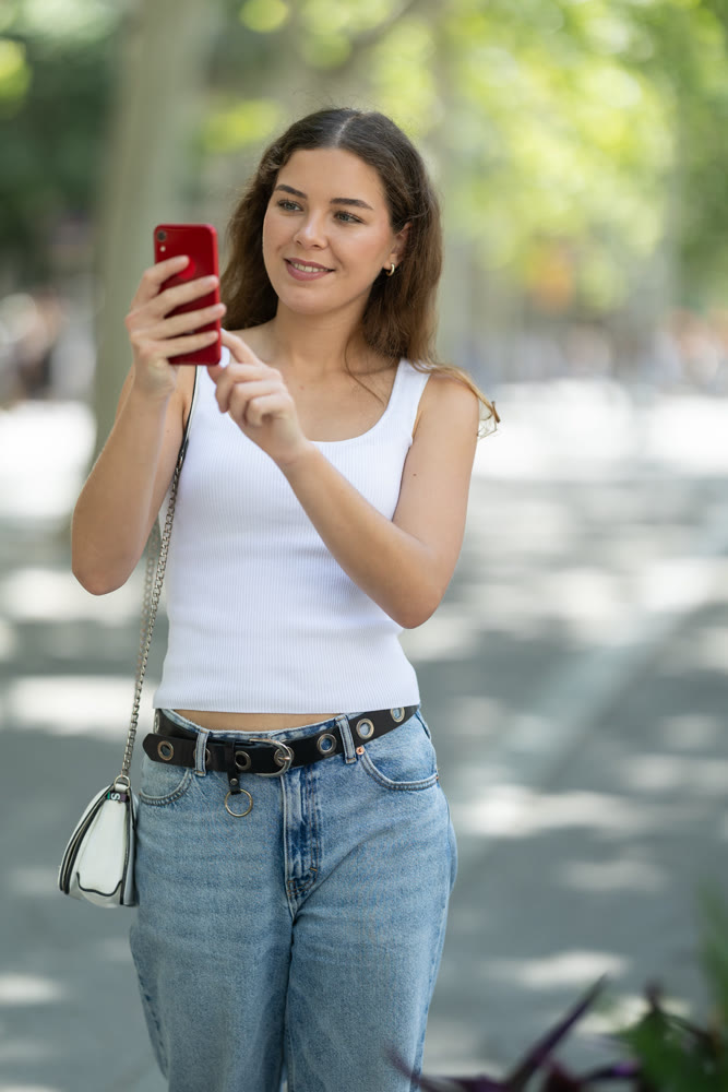Girl with phone walks along sunlit street, photographs architectural objects