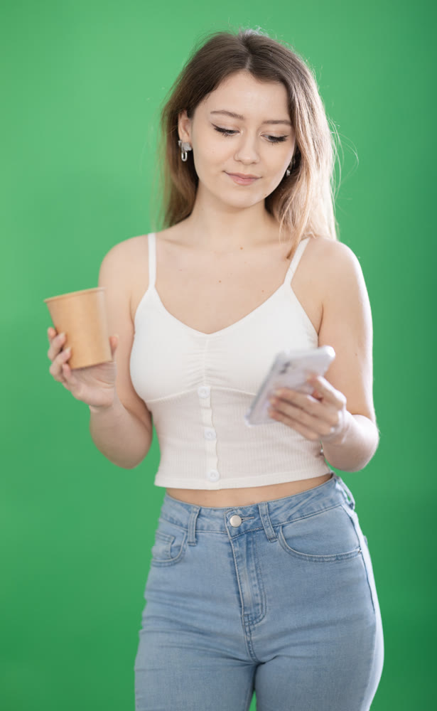 Woman standing on green background with smartphone and coffee in hands