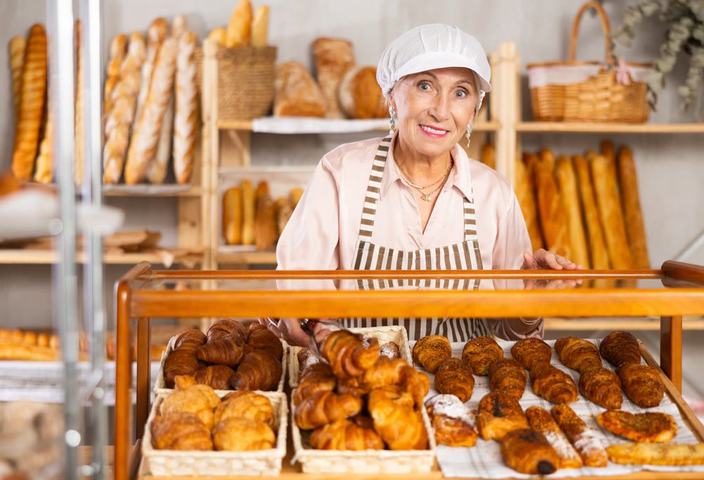 Senior woman employee puts croissants in window, arranges display of goods at bakery.