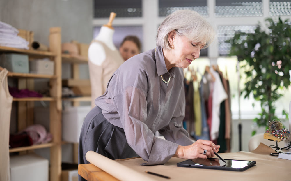 Elderly woman draws pattern on tablet female assistant sews on mannequin