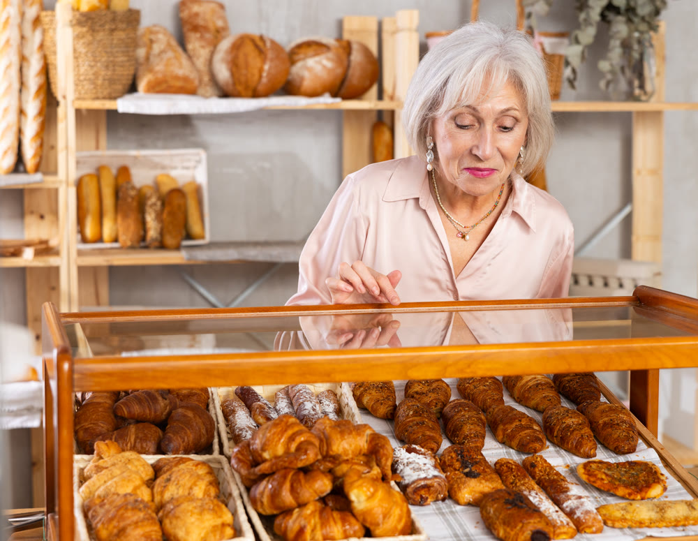 Mature European woman near window of bakery food court store examines assortment