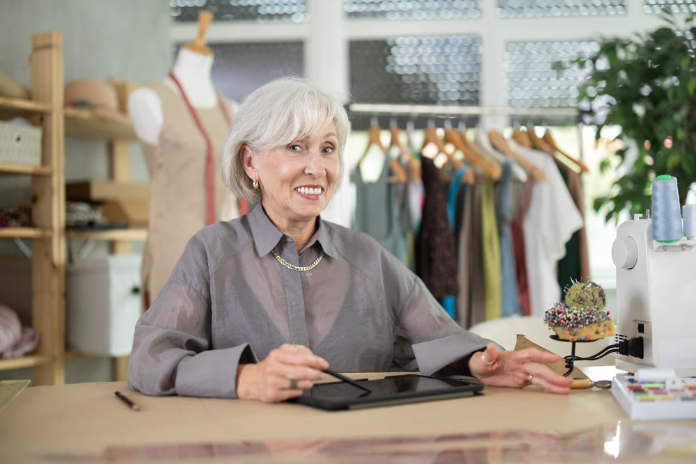 Positive mature female dressmaker drawing in tablet while sitting at table