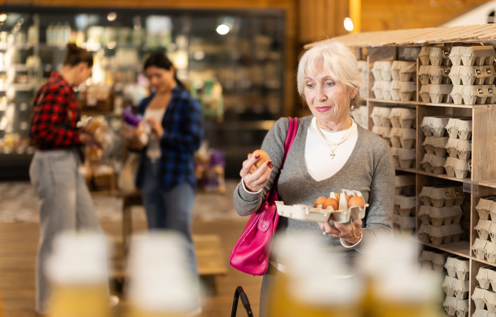 Elderly female customer selects and buys chicken eggs at grocery supermarket