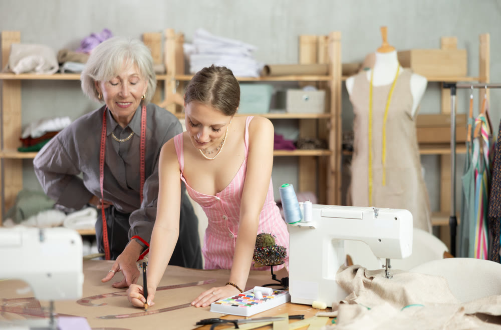 Elderly woman seamstress teaches young woman to draw pattern on paper