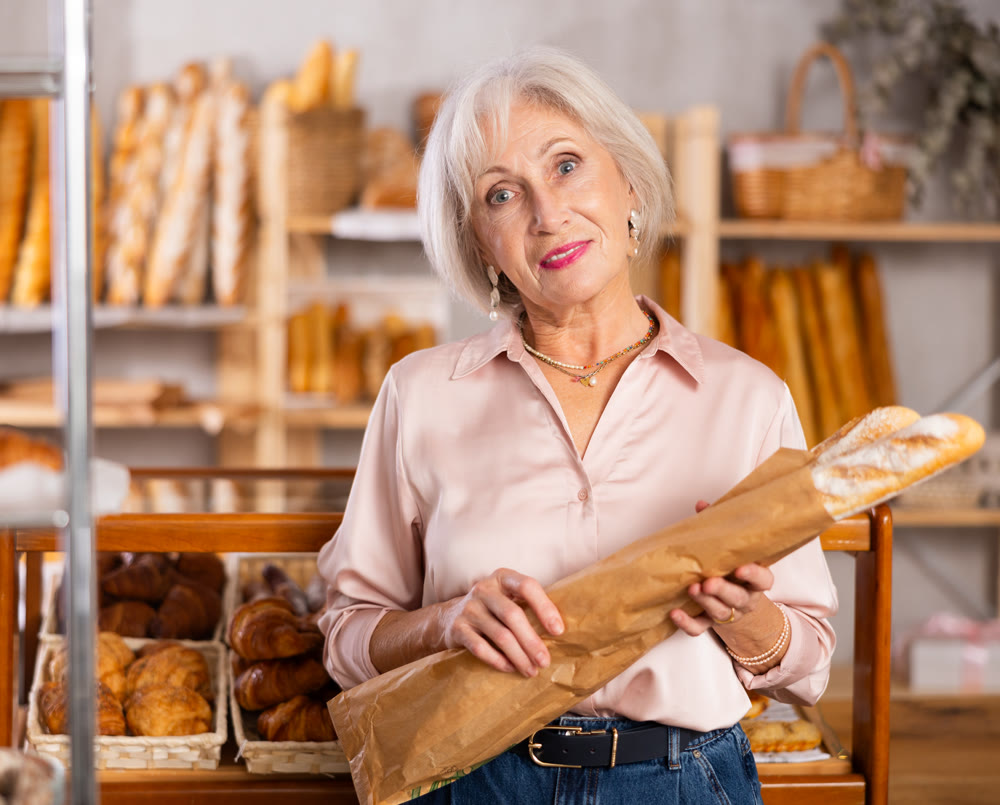 Elderly woman holds baguette wrapped in paper bag in bakery store