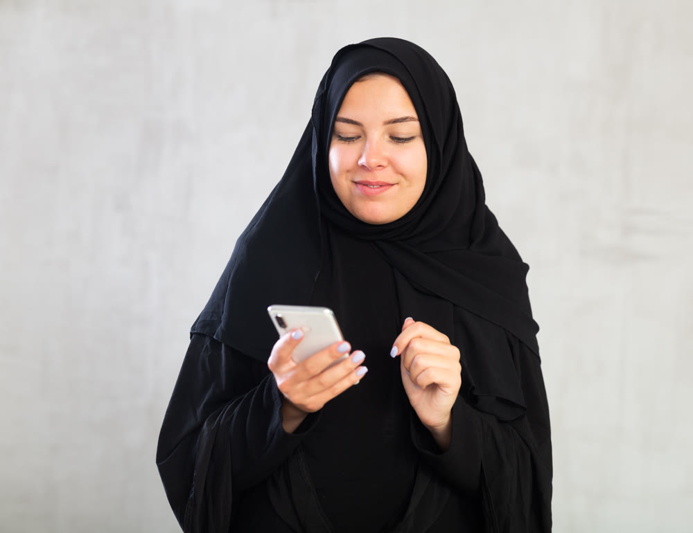 Smiling young woman dressed in traditional islamic clothes looking up something on her phone