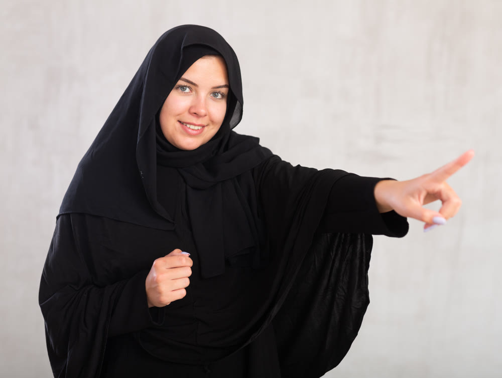 Cheerful young muslim woman dressed in black hijab pointing at side