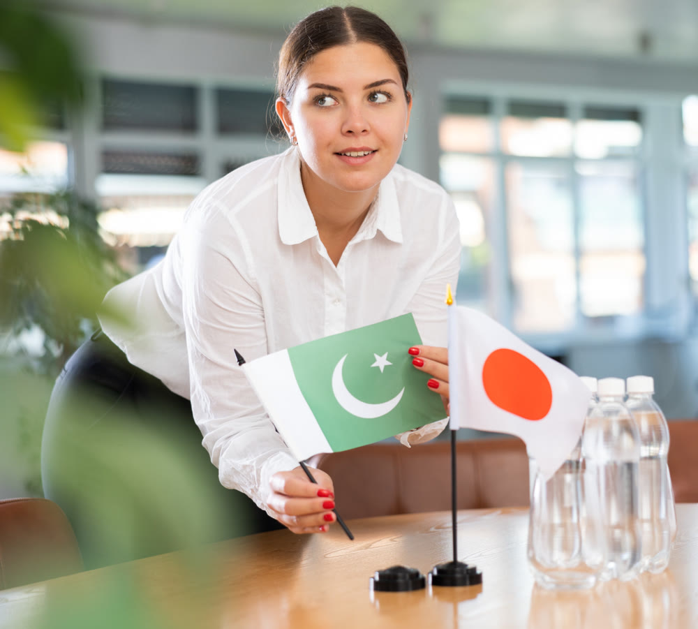 Positive young woman putting little flag of Pakistan on table next to the flag of Japan