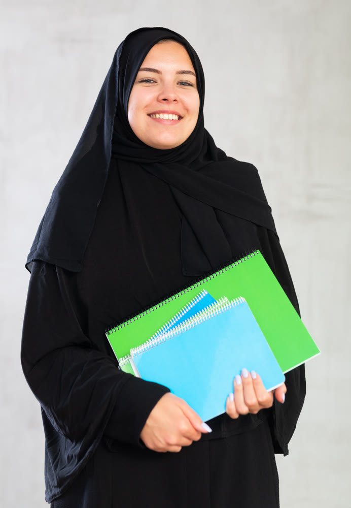 Smiling young female student dressed in traditional islamic clothes holding textbooks