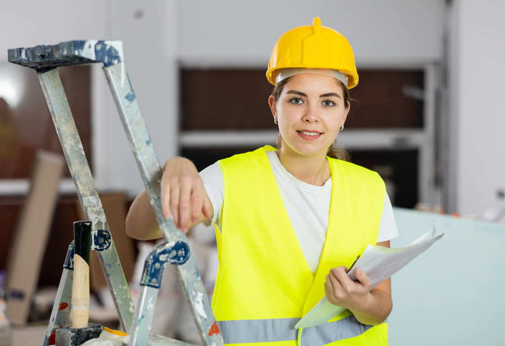 Woman foreman in a protective helmet and yellow vest checks the completed construction work on drawing