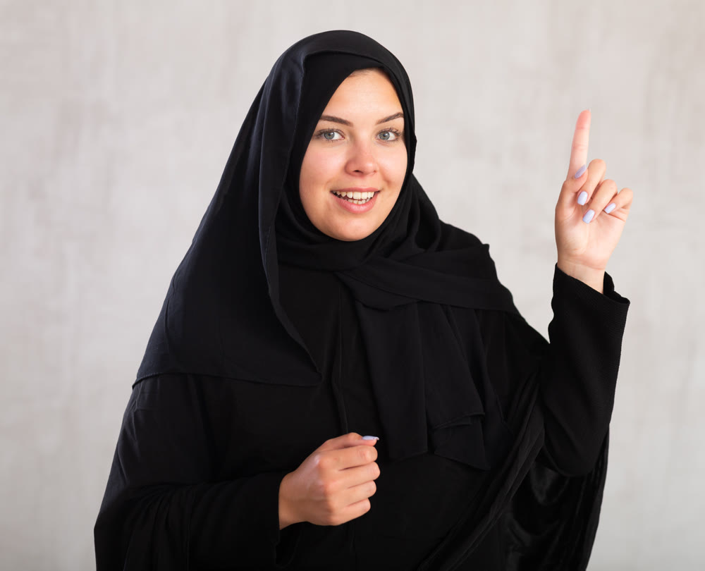 Cheerful young muslim woman dressed in black hijab pointing at side