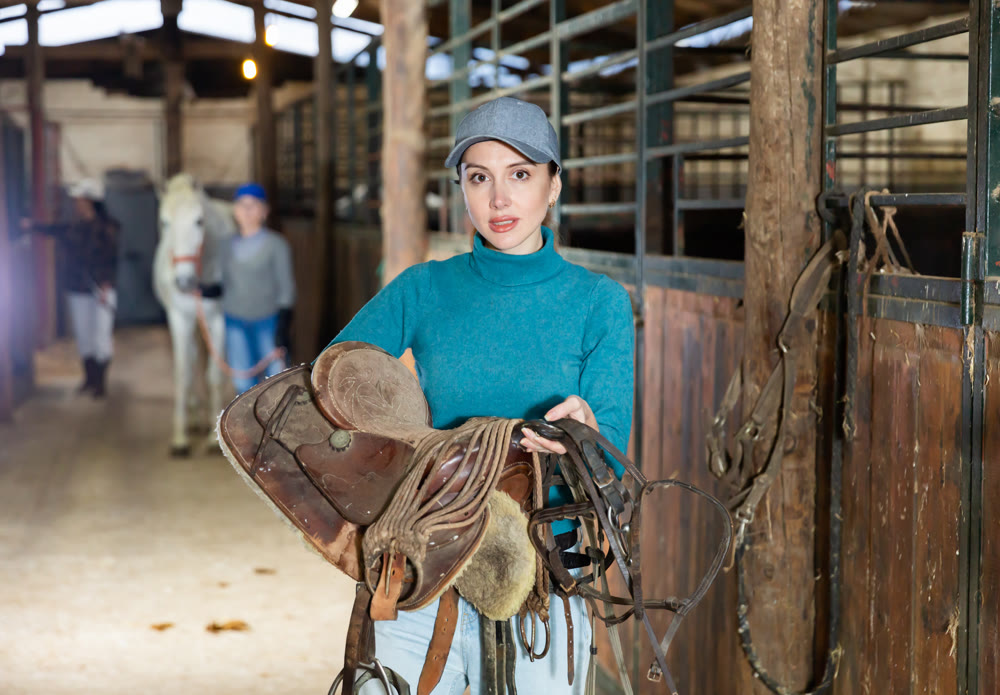Woman jockey with saddle and bridle in her hands in stable
