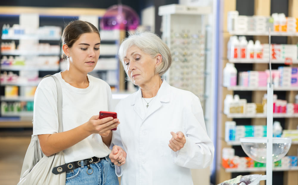 Female shopper shows name of drug on smartphone screen. Female elderly pharmacist helps to find medicine in pharmacy