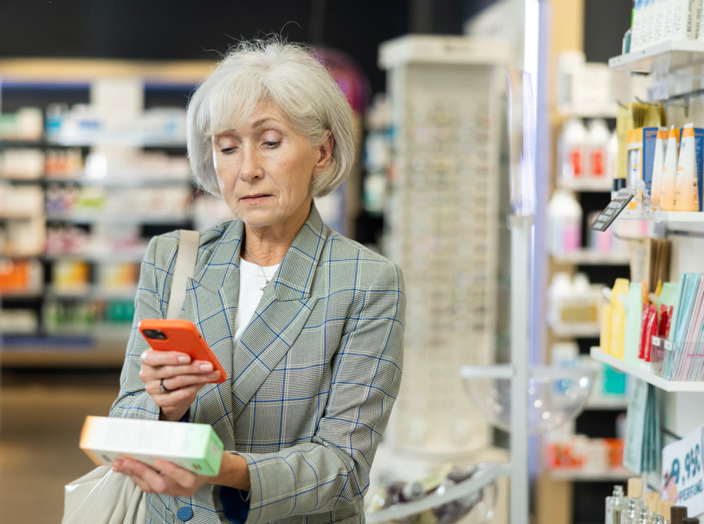 Mature woman scanning barcode on ointment package with her phone in pharmacy. Searching for information via Internet