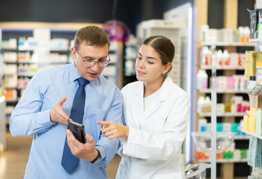 Friendly female pharmacist helping adult man with electronic prescription in phone choosing medical supplies at drugstore