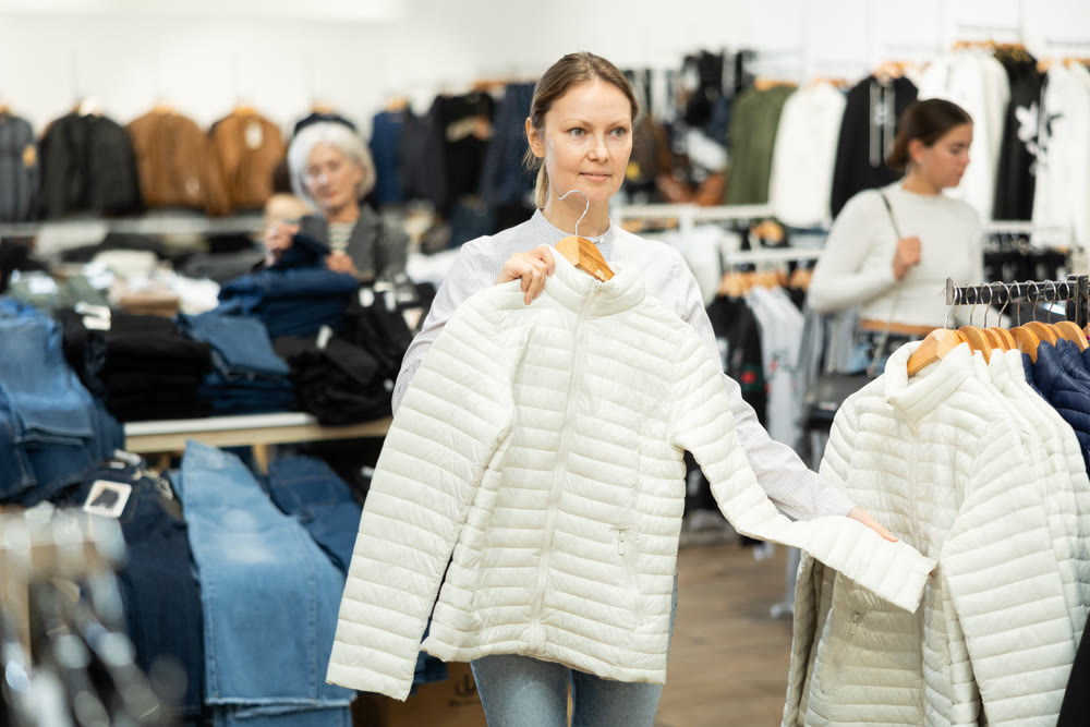 Positive woman chooses white winter down jacket in clothing store