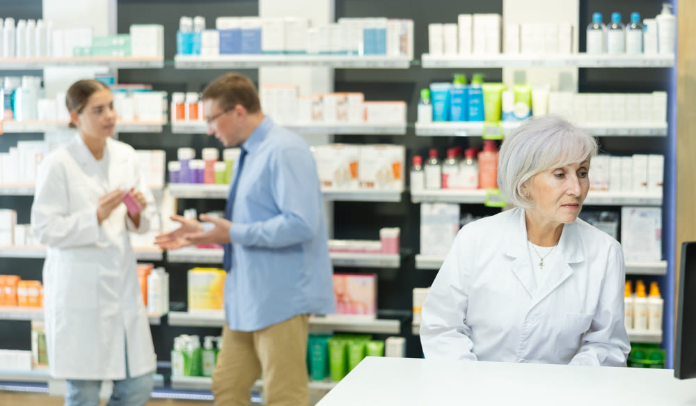 Portrait of an elderly friendly female pharmacist in white coat against background of display case with medicines in pharmacy