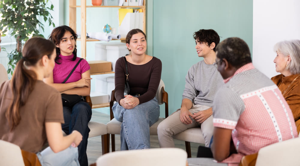 Male and female colleagues of various ages and nationalities talking sitting in circle on chairs in office lounge during break