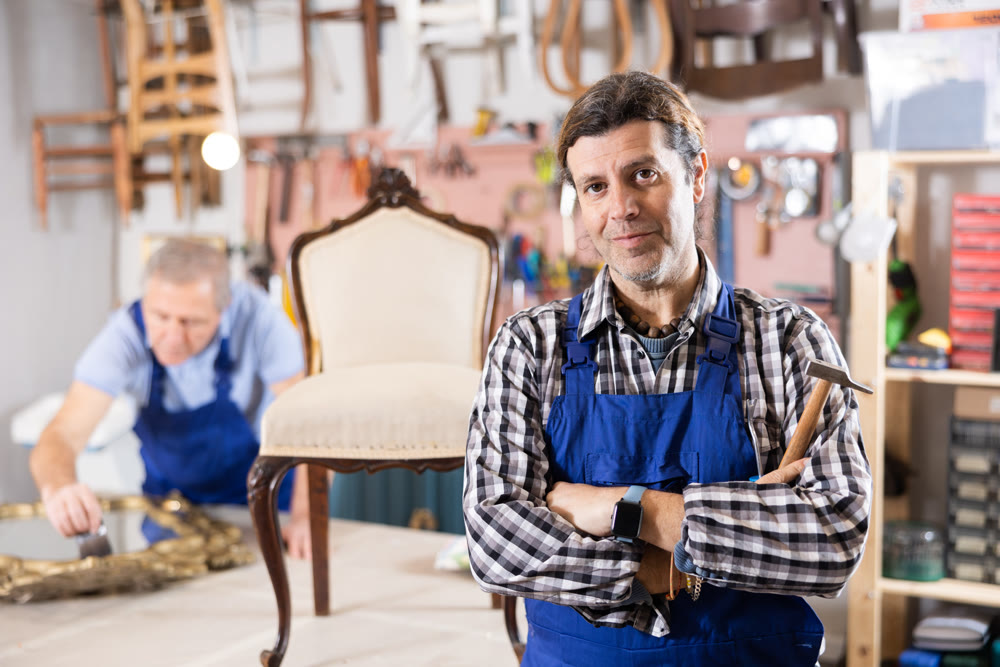 Portrait of middle-aged skillful repairman carpenter posing with crossed hands during workday in furniture woodwork studio