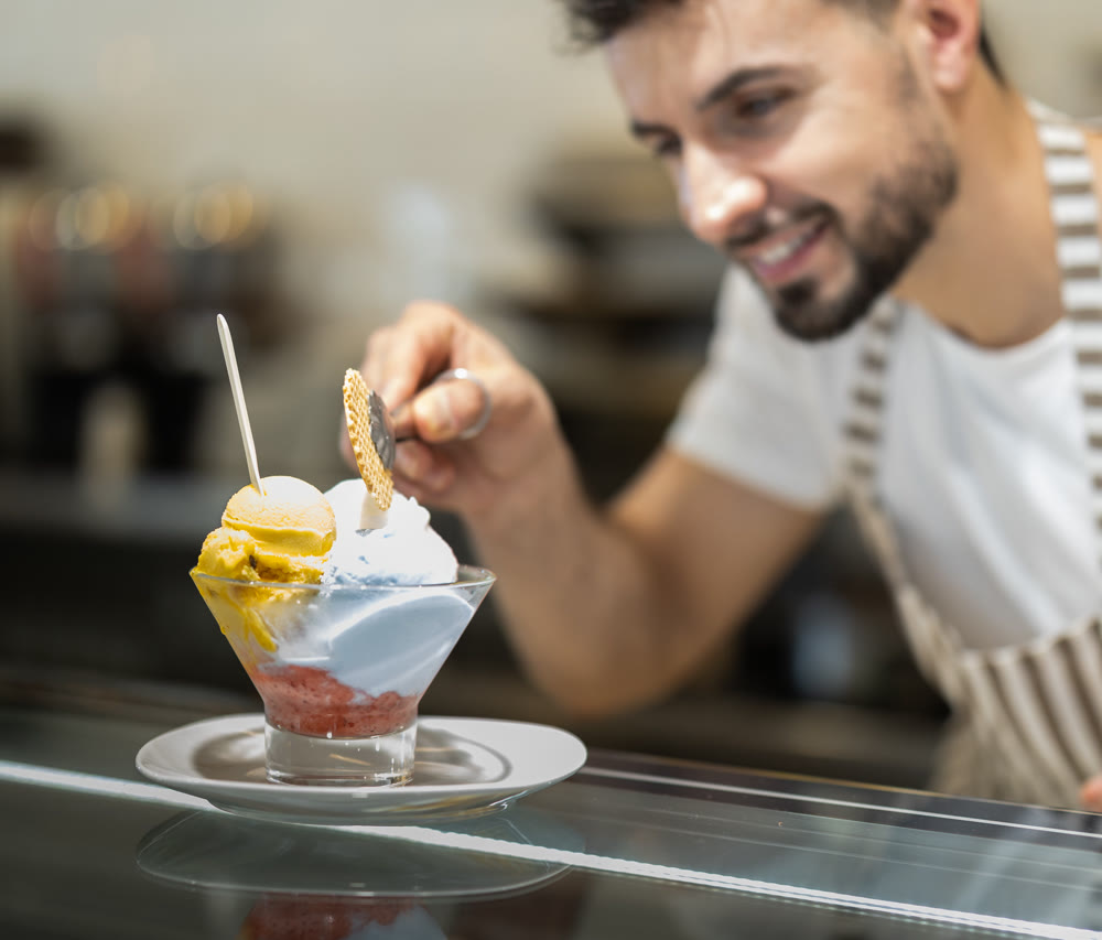 Positive man in an apron offers ice cream at dessert counter with an inviting smile