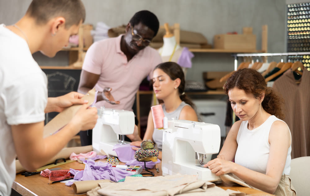 During lesson, African man teacher helps student sew, others work with fabric, blank, dress patterns