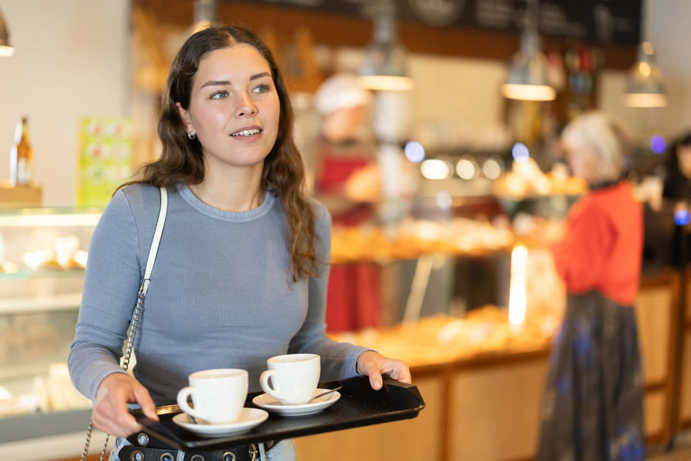 Girl visitor in self-service cafeteria carries tray with two cups of coffee.
