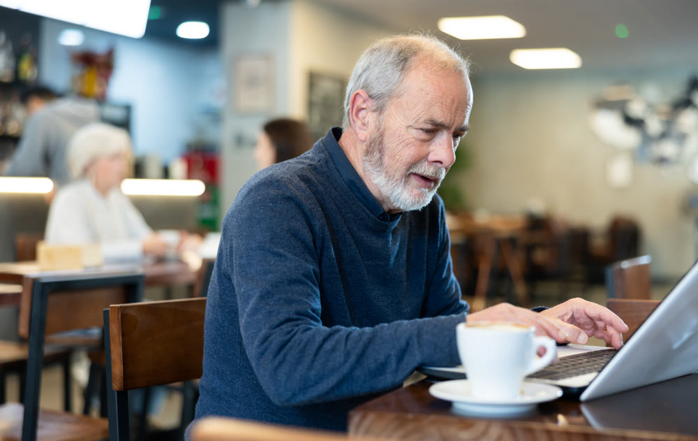 Older man is working on a laptop in a cozy cafe and drinking coffee