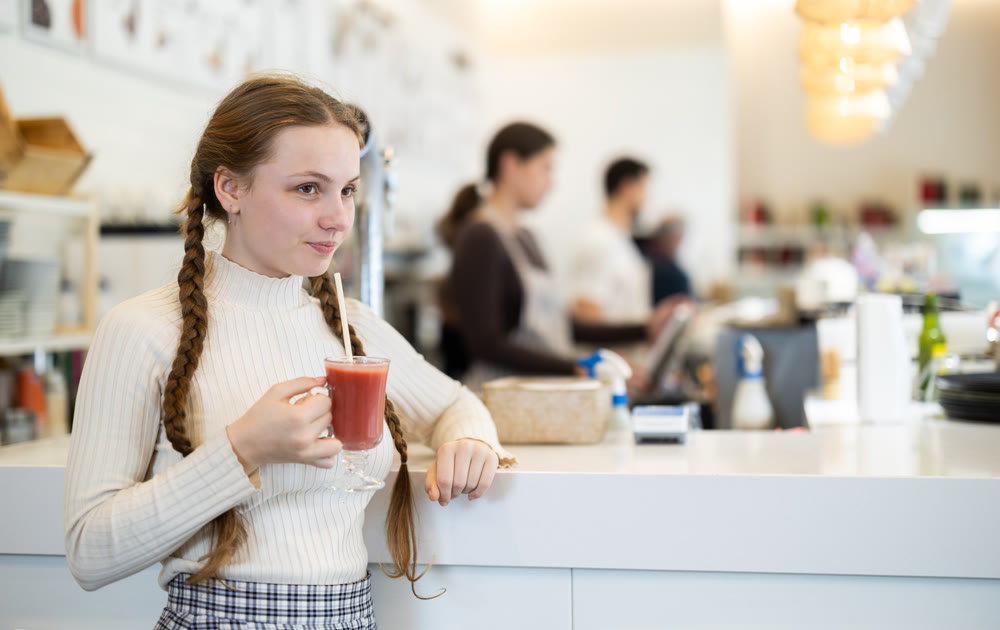 Teenage girl standing near the counter and drinking tomato juice