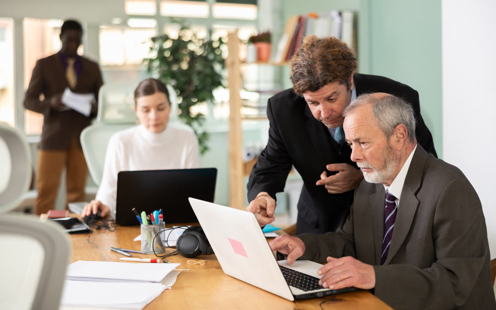 Middle aged man helping elderly employee with program on laptop
