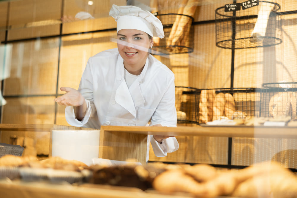 Female baker in a white coat and chef hat invites you to buy fresh bread and buns