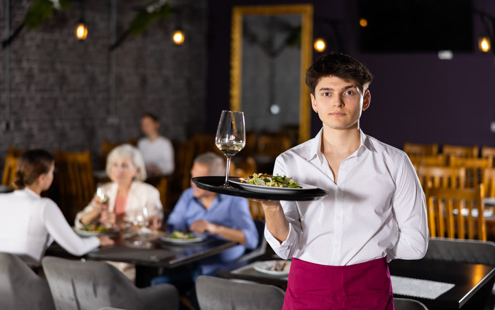 Young waiter guy is standing with order tray in spacious guest room in restaurant.